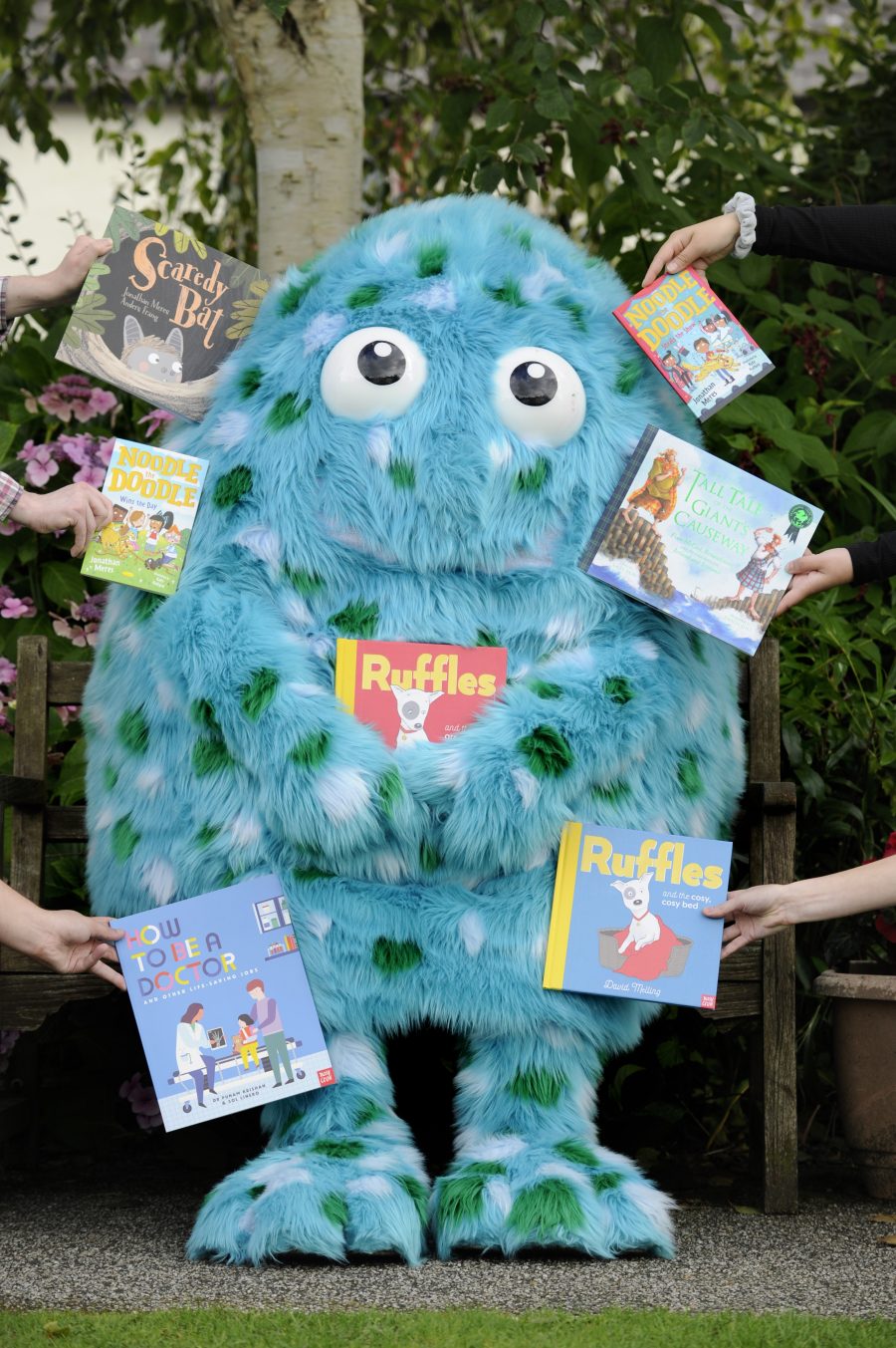 Bigwig, the Children's Book Festival mascot stands in the Wigtown Gardens holding an open book. Arms holding books are surrounding Bigwig.