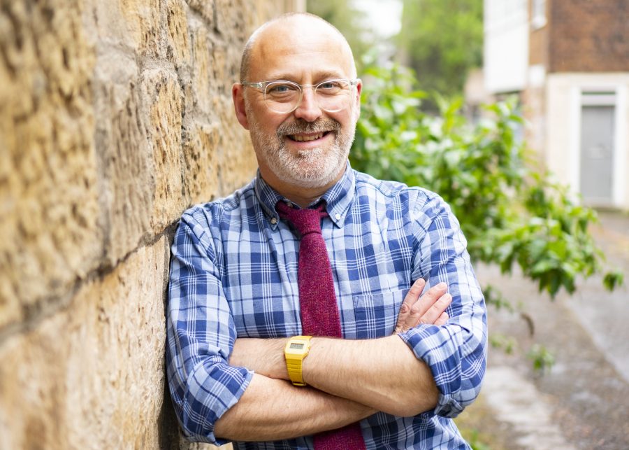 Headshot of Justin Davies standing against a wall with shrubs and a house behind him.