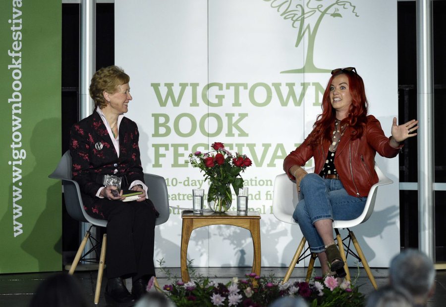 Hannah Jackson sitting on stage at her event at Wigtown Book Festival. She is talking animatedly to chairperson Polly Pullar. A table with flowers and water glasses is in between them.