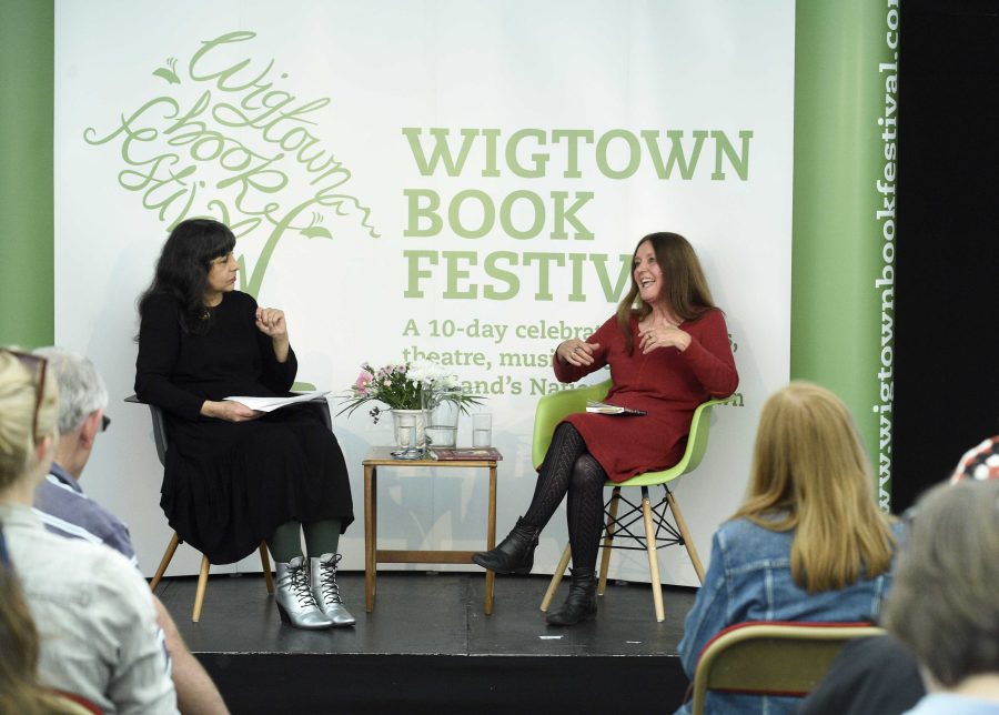 Gerda Stevenson sitting on stage with chairperson Lee Randall at Wigtown Book Festival. A table containing flowers, water and glasses is in between them. The audience sit before them listening.