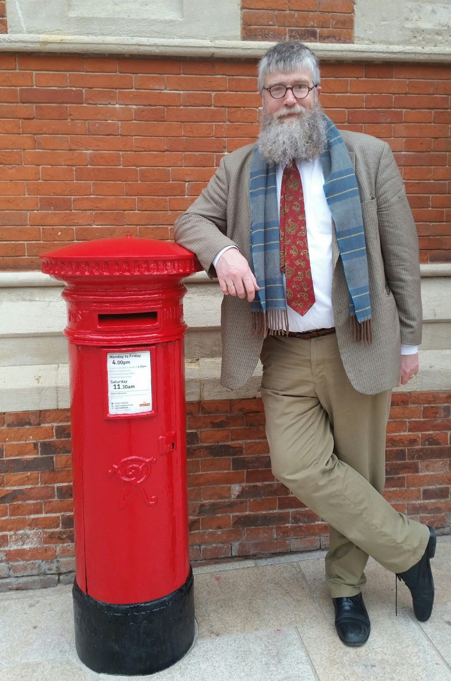 Philip Ardach standing in front of a red brick wall resting his arm on a red post box.