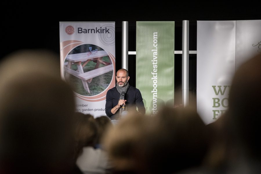 Andrew Cotter sitting on stage at his event at Wigtown Book Festival. He is holding a microphone as the audience sits in front of him.