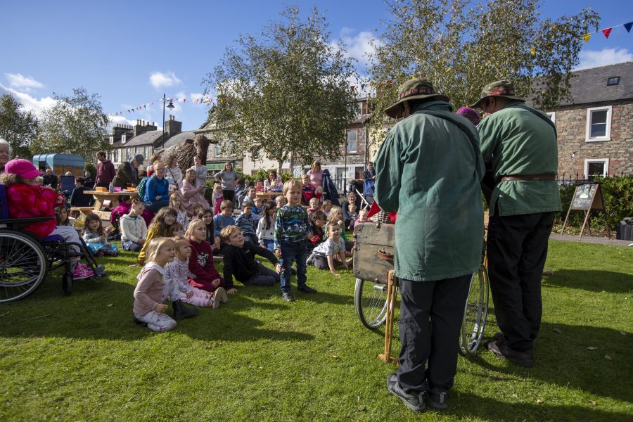 At an outdoor Children's Book Festival event a crowd sit in the gardens engaging with two entertainers in green jackets and brown hats. The terraced houses of Main Street in the background.