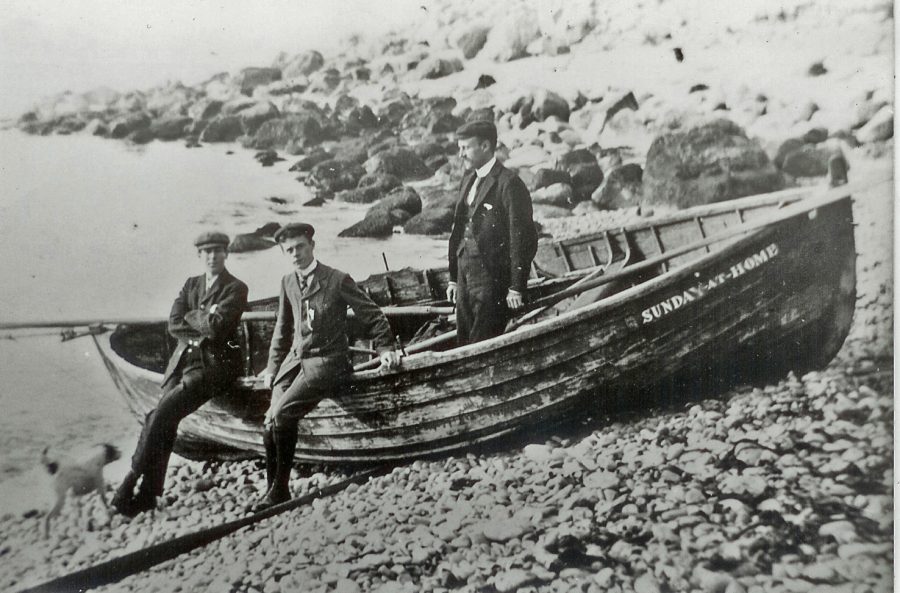 Victorian black and white photo of rowing boat on a stony beach, three men in suits present looking towards the camera. Small dog at one man's feet.