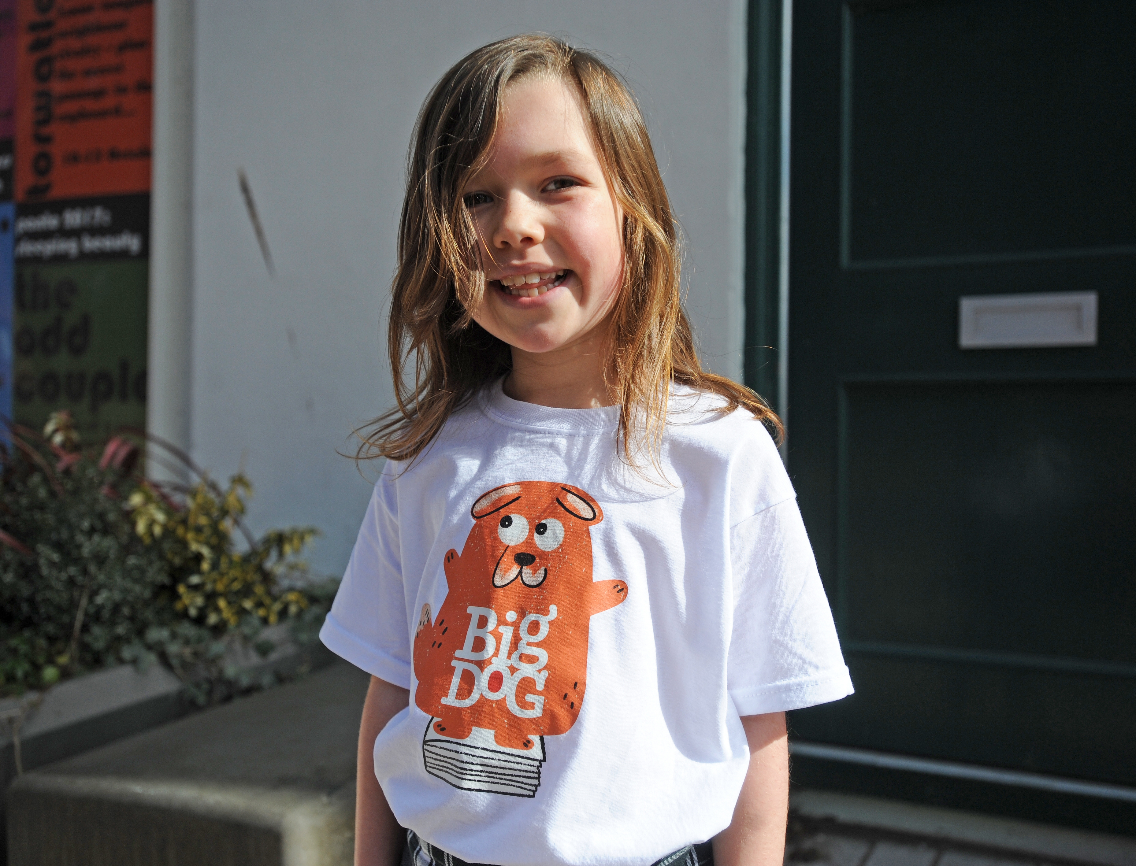 A young child standing smiling in front of a green door wearing a Big Dog t-shirt.