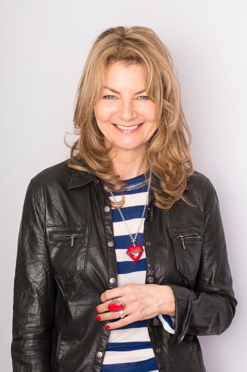 Author jo Caulfield stands smiling wearing a black jacket and blue and white striped t-shirt.