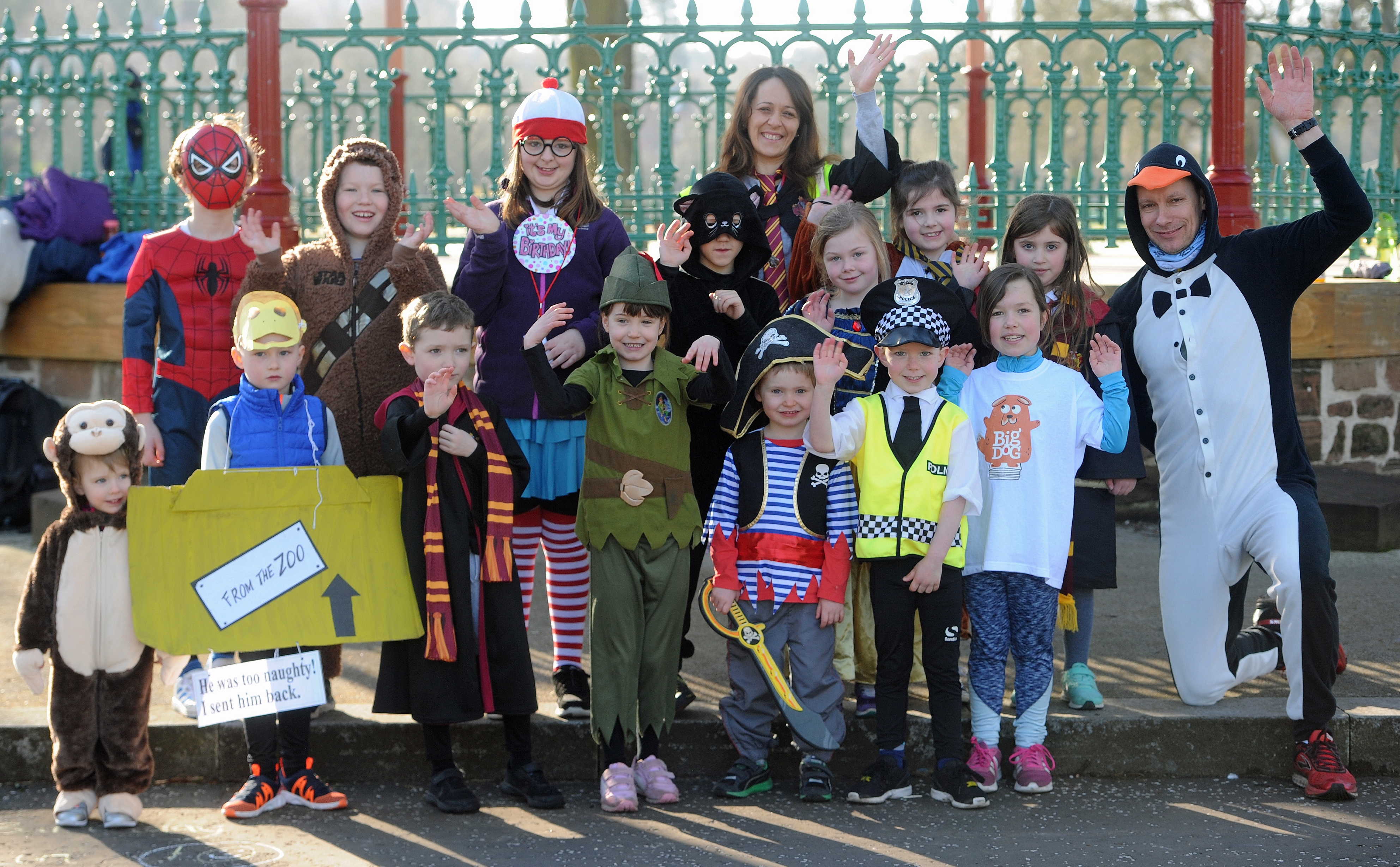 A group of children dressed up as various book characters stand waving and smiling at a Children's Book Festival event. Green railings are behind them.