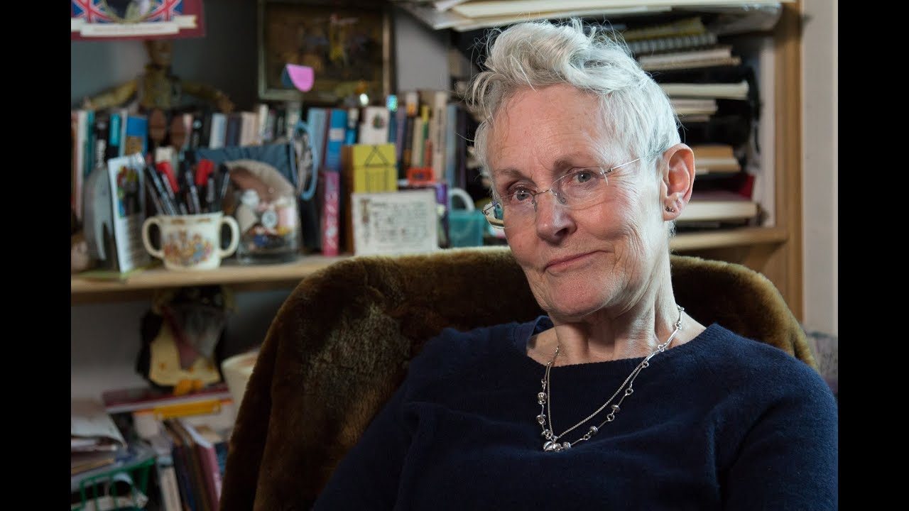 Author Vivian French  sitting in her office chair. Behind her are shelves full of  books, pens and  notebooks.