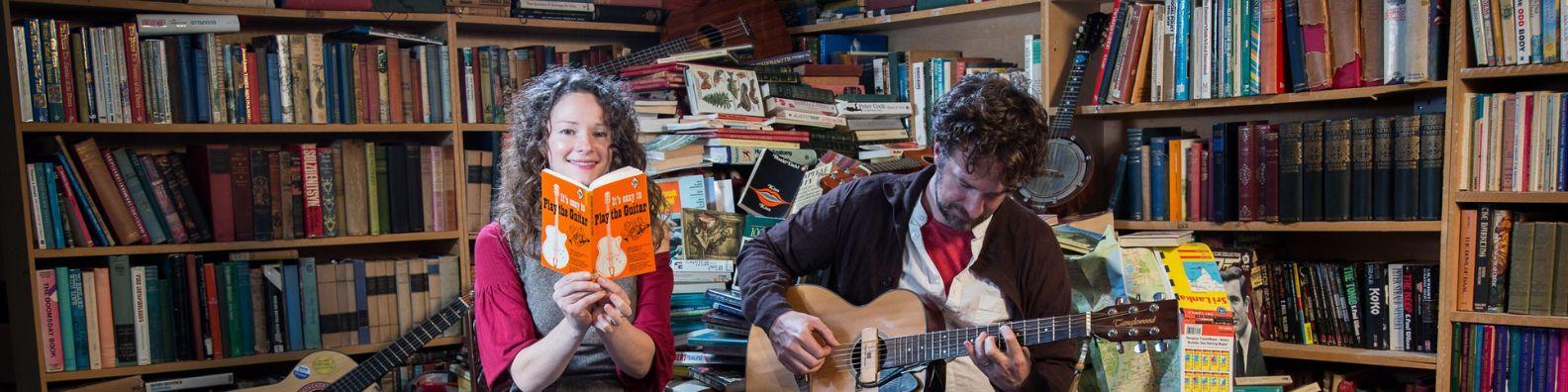 Bookshop band duo Beth and Ben Please sitting in a bookshop surrounded by books. Beth holds a book while Ben plays his guitar.